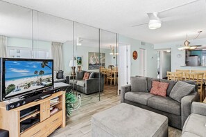 Ein modernes Wohnzimmer mit Flachbildfernseher, grauen Sofas, einer Holz-Entertainmentanlage und einem Essbereich, der durch eine Glasscheibe sichtbar ist.