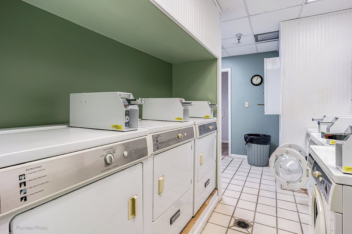 Une buanderie équipée de plusieurs machines à laver et de plusieurs sèche-linge, d’une poubelle et d’une horloge murale.
