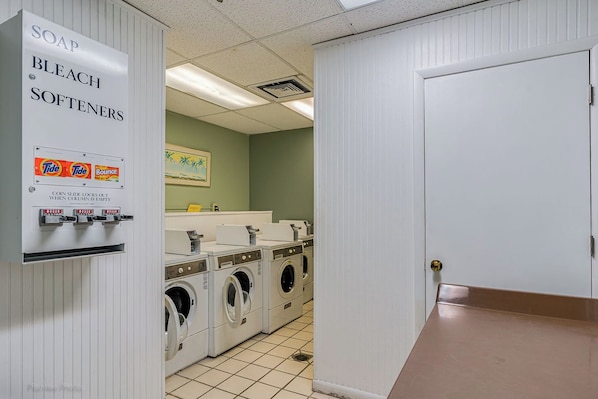 Ein Waschraum mit Münzwaschmaschine und Trockner, Spender für Waschmittel, Bleichmittel und Weichspüler sowie eine Tür rechts.