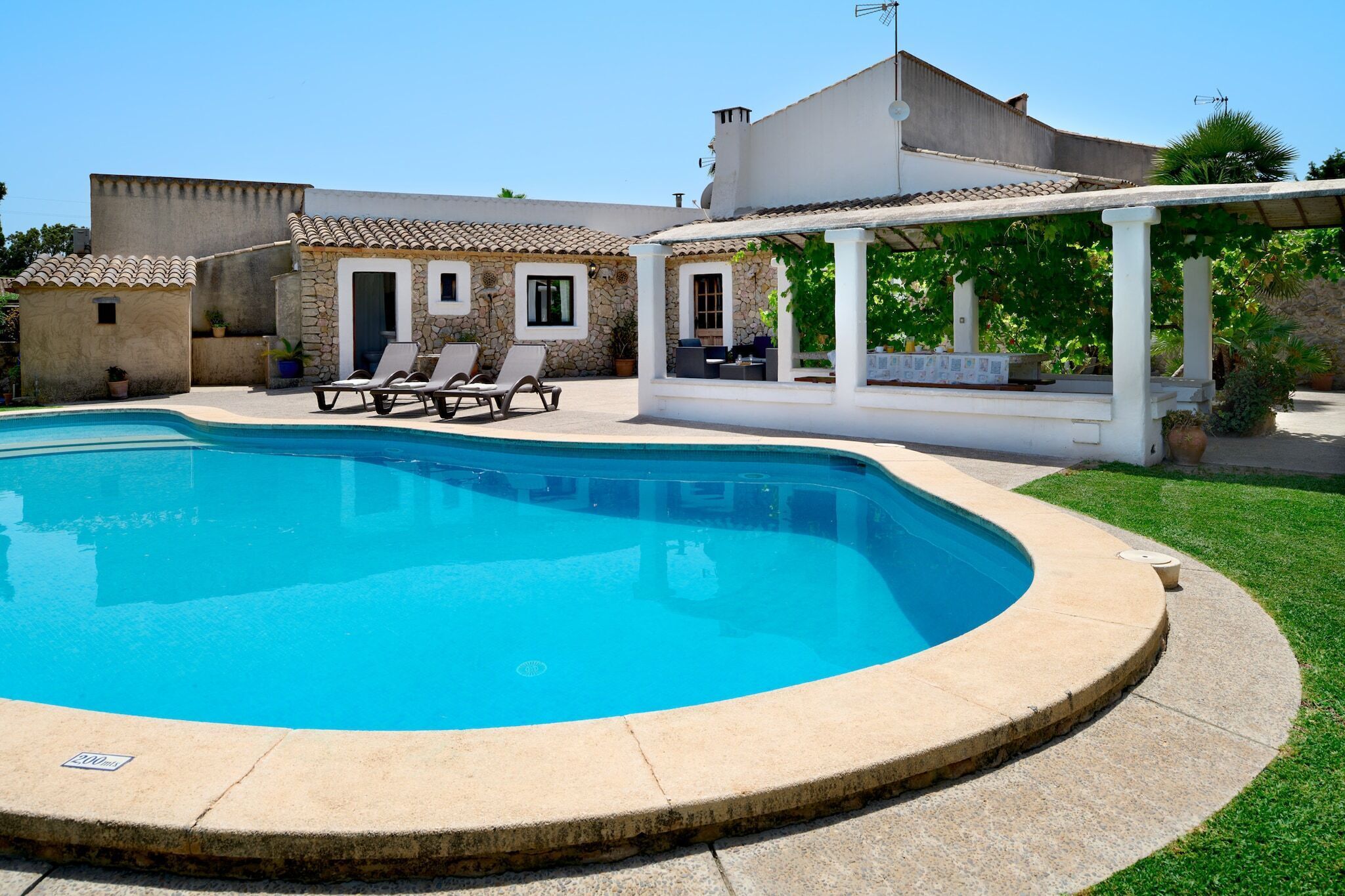 A swimming pool with a stone edge, surrounded by a grassy lawn and a white building with a tiled roof.