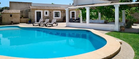 A swimming pool with a stone edge, surrounded by a grassy lawn and a white building with a tiled roof.