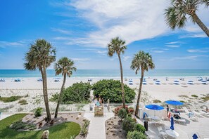 A beachside view with palm trees, a pathway leading to beach umbrellas, and a clear sky. - Gulf Front Getaway! Sand Castle I 307 (Indian Shores)