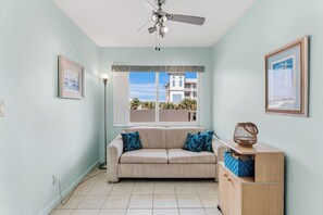 A room with a beige sofa, a ceiling fan, a window with a view of buildings, and a wooden cabinet. - Gulf Front Getaway! Sand Castle I 307 (Indian Shores)