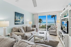 A living room with a sofa, coffee table, and TV, a balcony with a view of the sea, and a painting of sailboats. - Gulf Front Getaway! Sand Castle I 307 (Indian Shores)