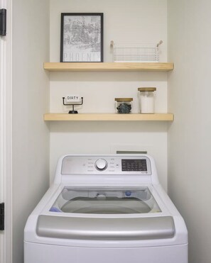 A laundry area with a front-loading washing machine, two wooden shelves above it, and a framed map on the wall. - Desert Bliss Villa - 3 bedroom Heated Pool, Putting Green, Pool Table & Games (Phoenix)