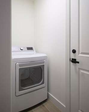 A compact, white, front-loading washer and dryer unit in a small laundry room with a door to the right. - Desert Bliss Villa - 3 bedroom Heated Pool, Putting Green, Pool Table & Games (Phoenix)