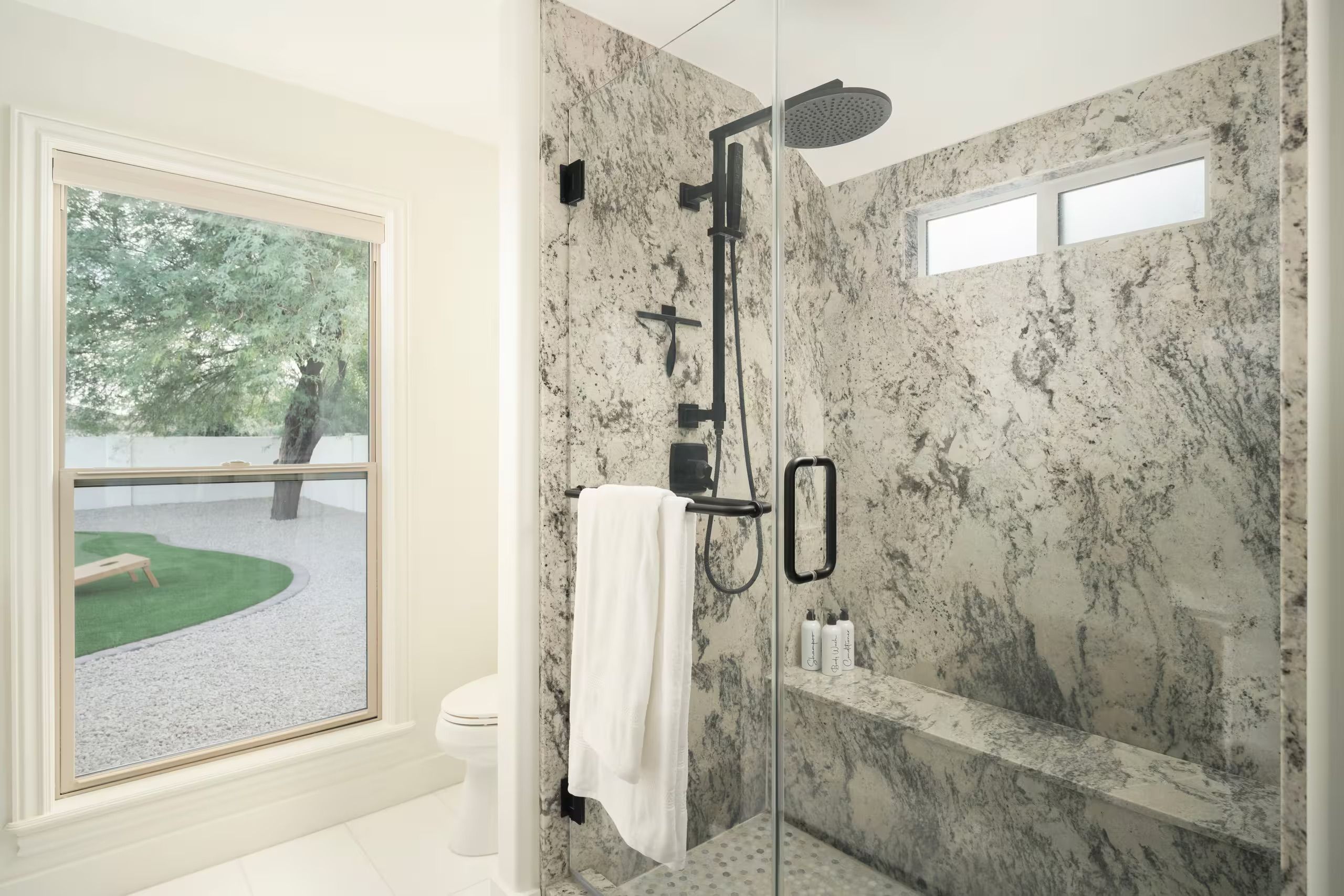 A modern bathroom with a glass-enclosed shower, marble walls, a toilet, and a window with a view of a garden.