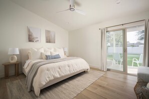 Ein Schlafzimmer mit einem Bett, einem Nachttisch, einer Lampe, einem Deckenventilator und Blick ins Freie durch eine Schiebetür.