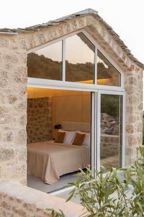 A bedroom with a stone exterior, a bed with a beige bedspread, and a view through a large window.