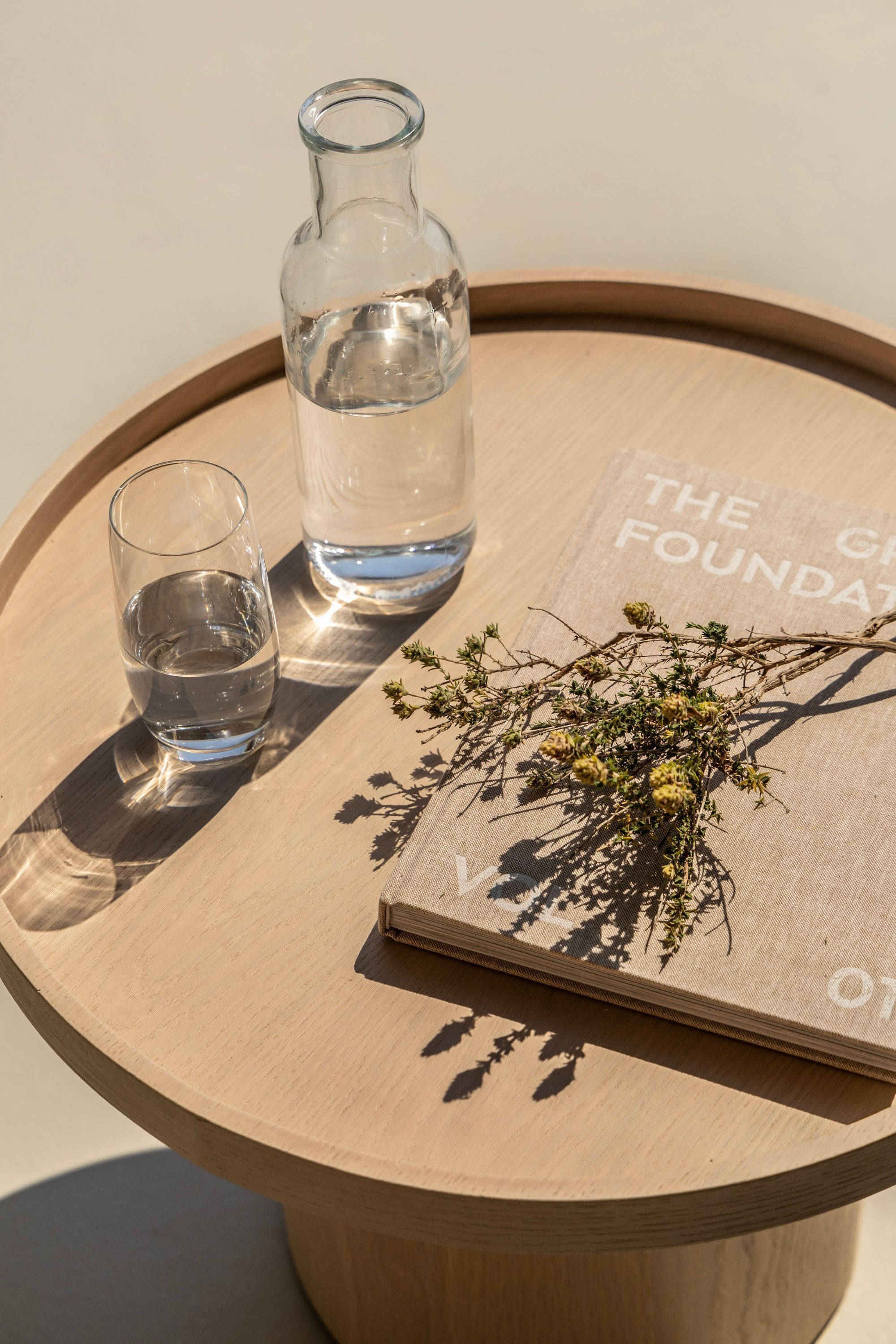 A round wooden table with a clear glass bottle and a glass of water, a small glass, and a decorative branch on a fabric surface.