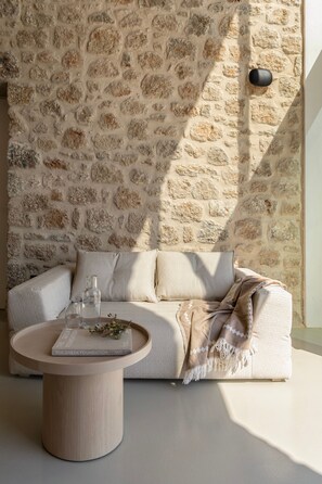 A room with a stone wall, a beige sofa, a round wooden coffee table, and a glass of water.
