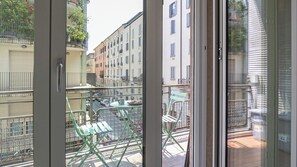 Un balcon avec une table et des chaises, donnant sur une rue bordée d’immeubles et de voitures garées.