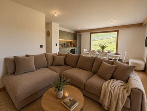 A modern living room with a large L-shaped sectional sofa, a coffee table with a book and a plant, and a dining area with a table set for four. - Ski aux pieds | Piscine, spa & salle de sport (Alpe d'Huez)
