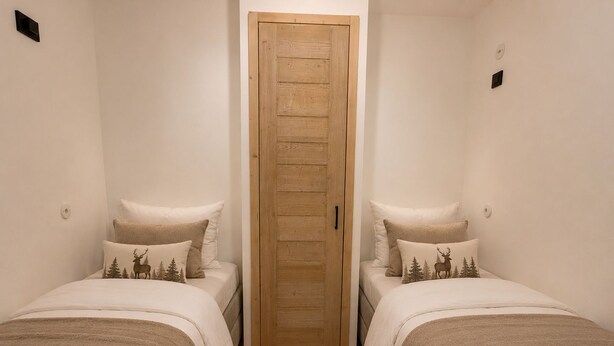 Ein Zimmer mit zwei Betten, jeweils mit Kopfteil und Kissen mit einem Motiv aus Hirschen und Bäumen. Eine Holztür befindet sich mittig zwischen den Betten.