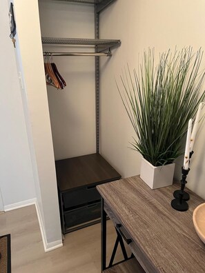 Ein kleiner Raum mit einem Holztisch, einer Topfpflanze, einem Metallregal und einem Kleiderständer.