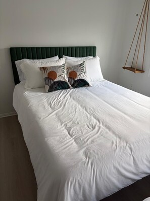 En snyggt bäddad säng med vita sängkläder och dekorativa kuddar, en grön klädd sänggavel och en trägungstol som hänger från taket.