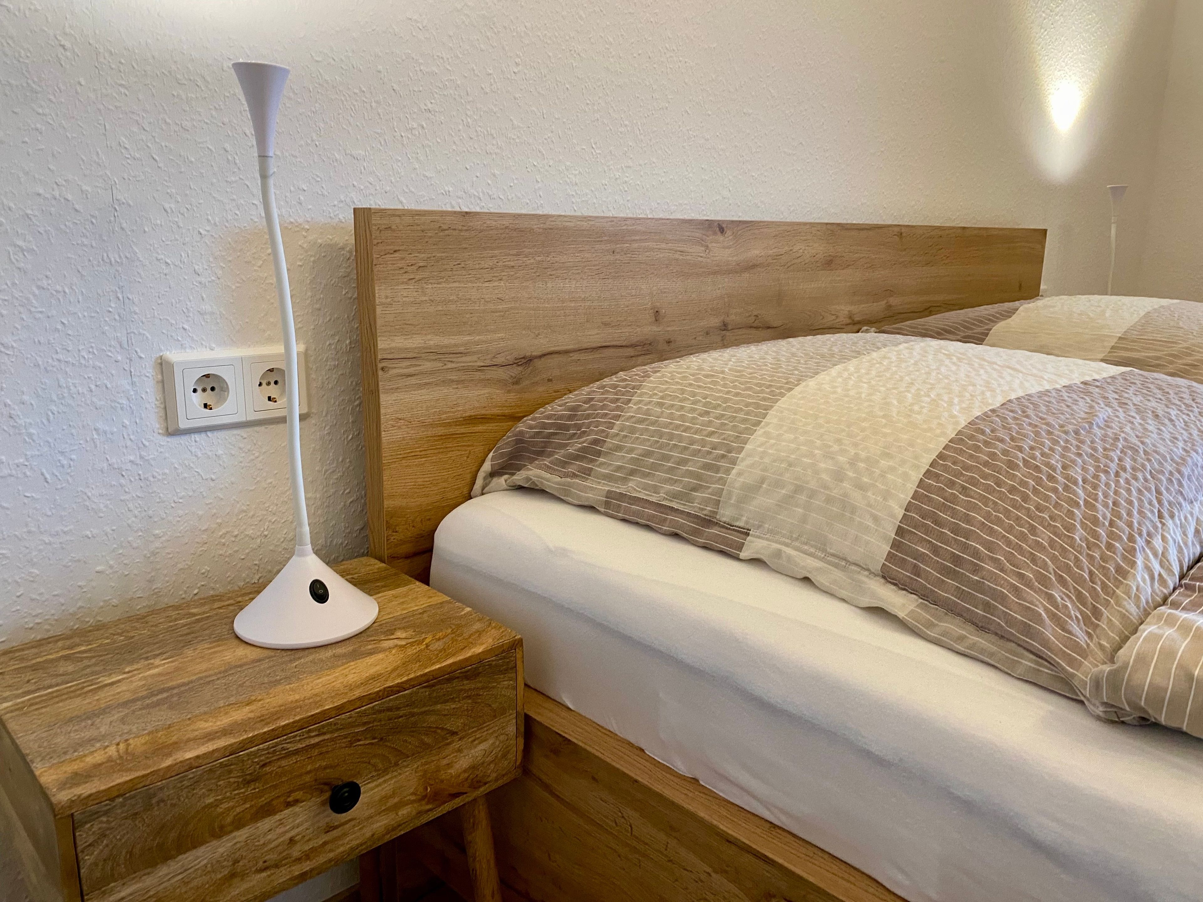 Ein Nachttisch aus Holz mit einer Lampe, ein Bett mit einer gestreiften Decke und eine Steckdose.