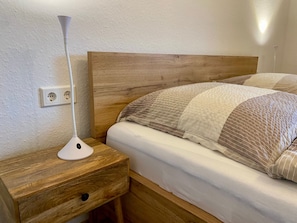 A wooden bedside table with a lamp, a bed with a striped blanket, and a wall socket.