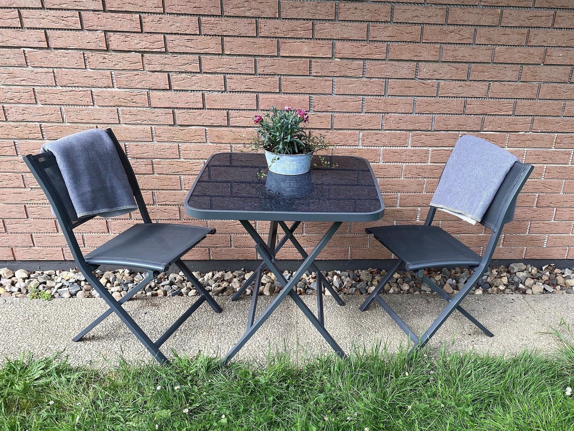 Un pequeño espacio al aire libre con dos sillas y una mesa, frente a un muro de ladrillo.