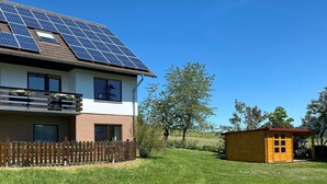 Ein zweistöckiges Haus mit Solaranlage auf dem Dach, einem Holzzaun und einer kleinen Holzbude im Garten.