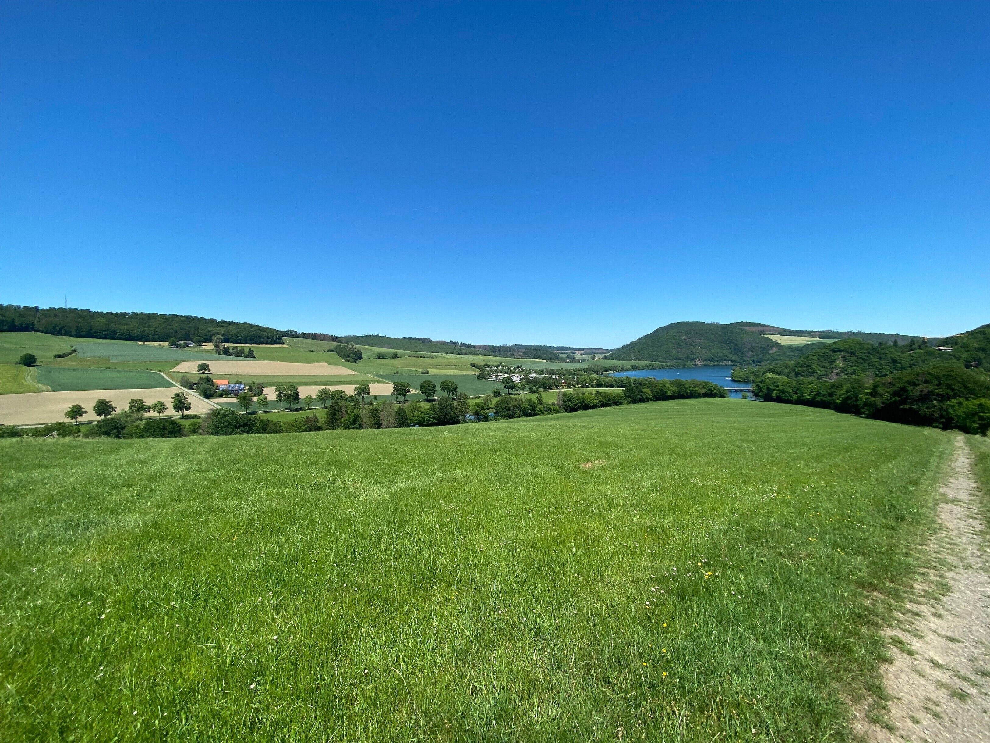 Un paysage rural avec un ciel dégagé, une rivière et un chemin de terre qui traverse un champ verdoyant.