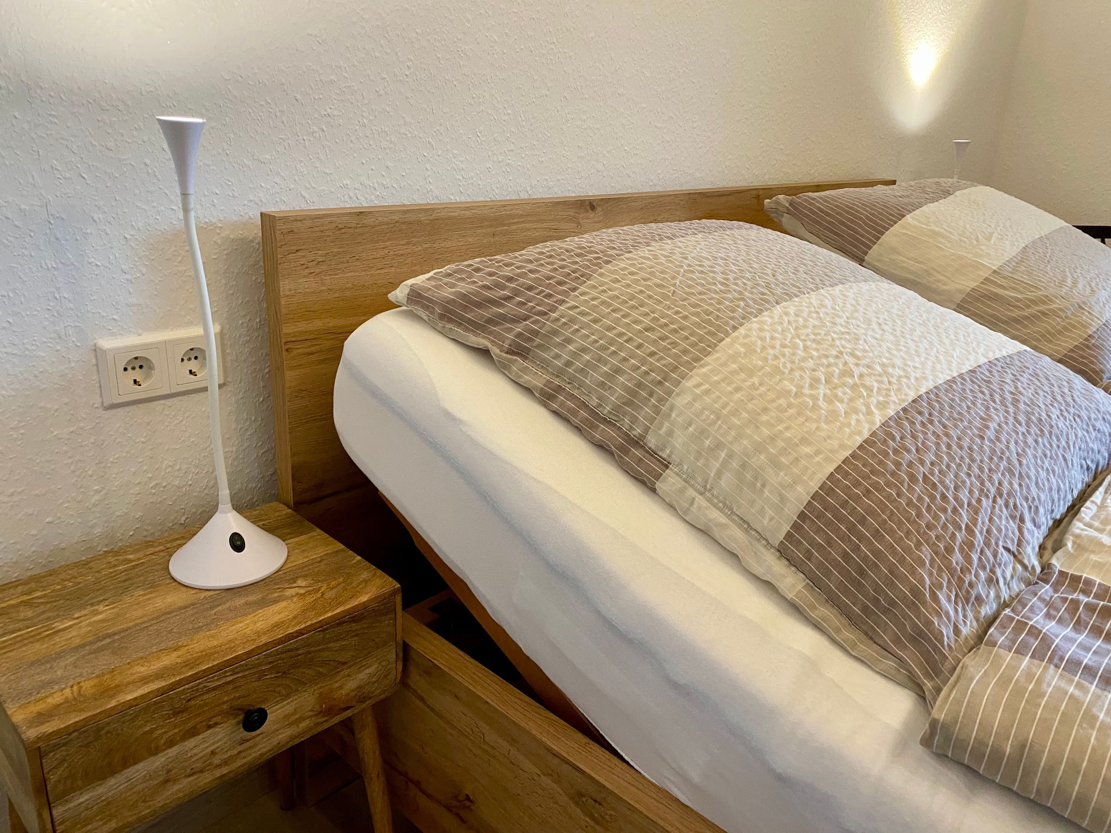 Ein ordentlich bezogenes Bett mit einem hölzernen Kopfteil, ein Nachttisch mit Lampe und eine Steckdose an der Wand.