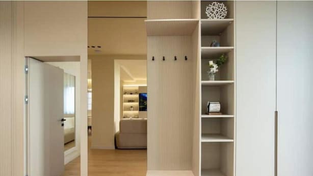 A modern interior with a wooden floor, a white shelving unit with decorative items, and a glimpse of a living area in the background.
