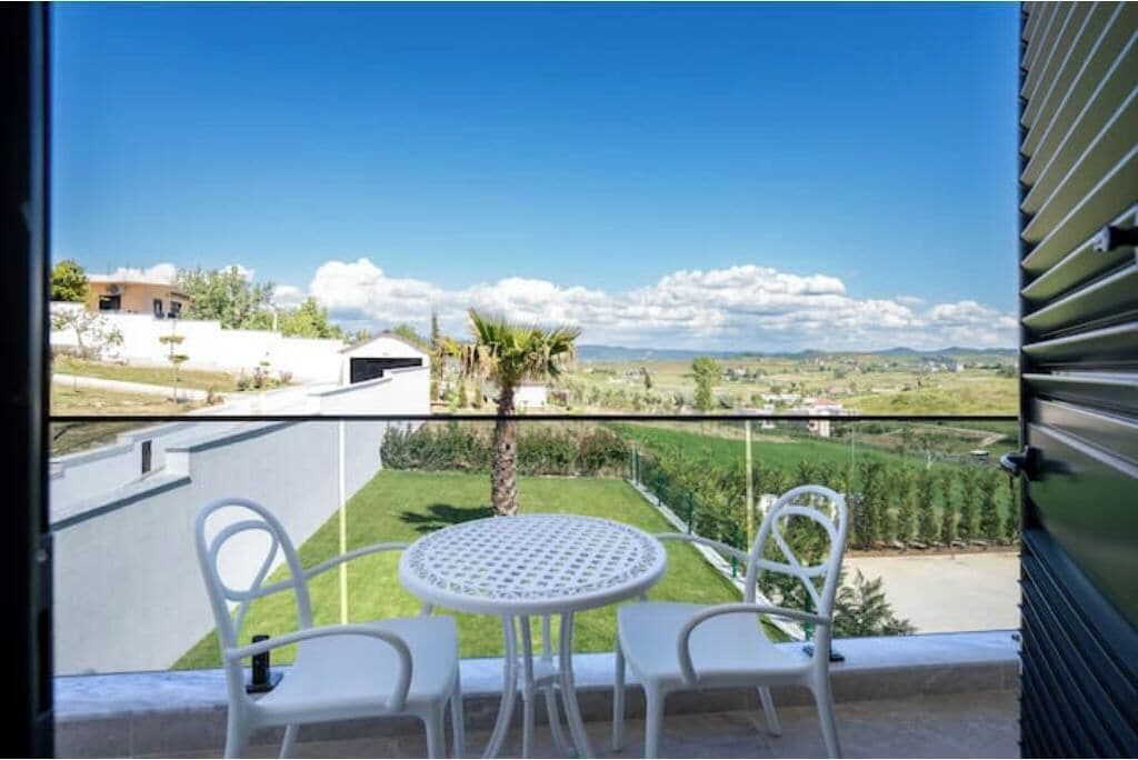 Un balcon avec une table et des chaises, donnant sur un paysage verdoyant et sur des bâtiments situés au loin.