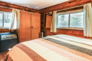 A cabin-style room with wooden walls, a bed with a striped blanket, a wooden wardrobe, and a window with curtains.