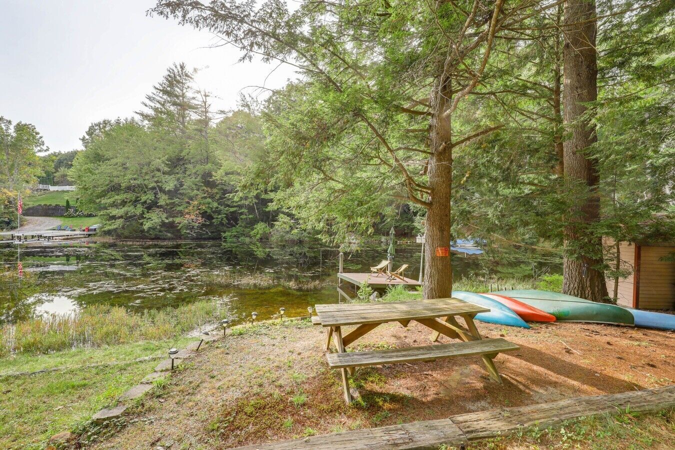 Una mesa de picnic y canoas junto a un lago, con árboles y un muelle al fondo.