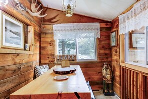 A rustic cabin interior with wooden walls, a dining table, a window with a view of trees, and a decorative antler on the wall.