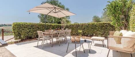 Un patio al aire libre con una mesa, sillas, un paraguas grande y una zona de estar con cojines.