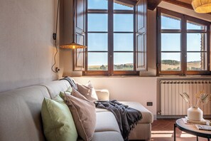 A cozy living room with a sofa, pillows, a table with books, and a view through large windows. - New Apartment in Tuscany with a Pool and Breathtaking Views (Asciano)