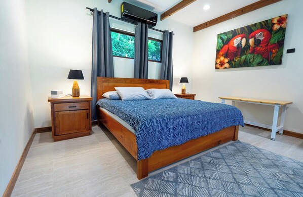 A bedroom with a wooden bed, two bedside tables with lamps, a window with curtains, a painting of parrots, and a bench. - Enjoyable 3-bedroom house with AC near Samara. (Samara)