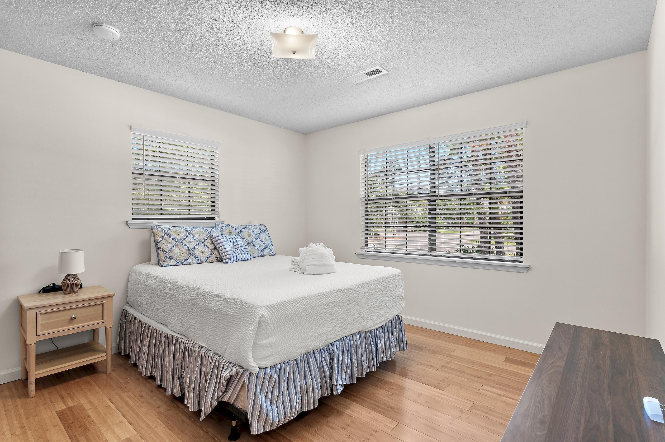 A bedroom with a bed, bedside table, lamp, and two windows with blinds.