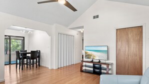 A modern living room with a flat-screen TV, wooden flooring, a ceiling fan, and a view of the beach through a window. - Ash at the Beach (Santa Rosa Beach)