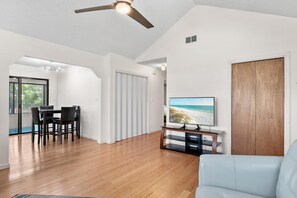 Ein modernes Wohnzimmer mit Flachbildfernseher, Holzboden, Deckenventilator und Blick auf den Strand durch ein Fenster.