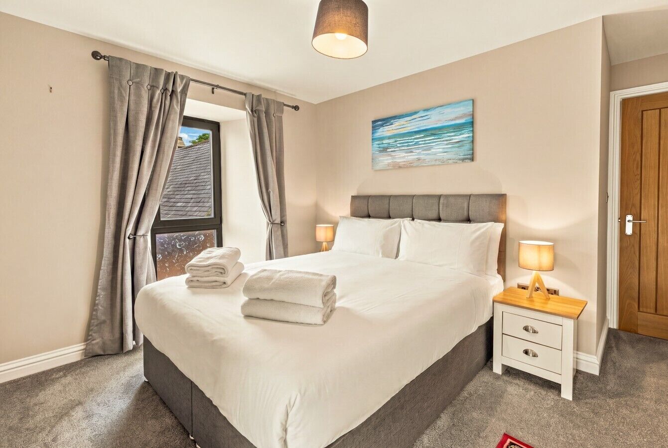 A neatly made bed with white linens and folded towels, a bedside table with a lamp, and a window with curtains.