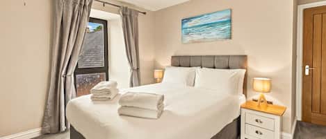 Una cama bien hecha con sábanas blancas y toallas dobladas, una mesita de noche con lámpara, y una ventana con cortinas.