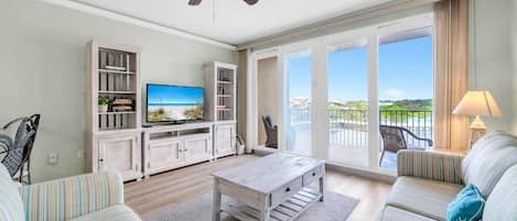 A spacious living room with a television, a ceiling fan, a sofa, a coffee table, and a balcony with a view.
