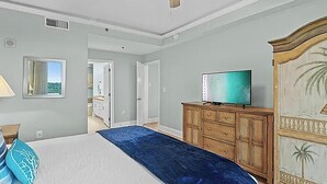 Une chambre à coucher avec un lit, un téléviseur posé sur une commode en bois, un ventilateur de plafond et une vue sur l’extérieur, visible à travers une porte ouverte.