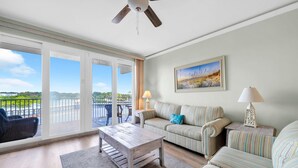 Un salon spacieux avec vue sur un plan d’eau, un ventilateur de plafond et un tableau accroché au mur.
