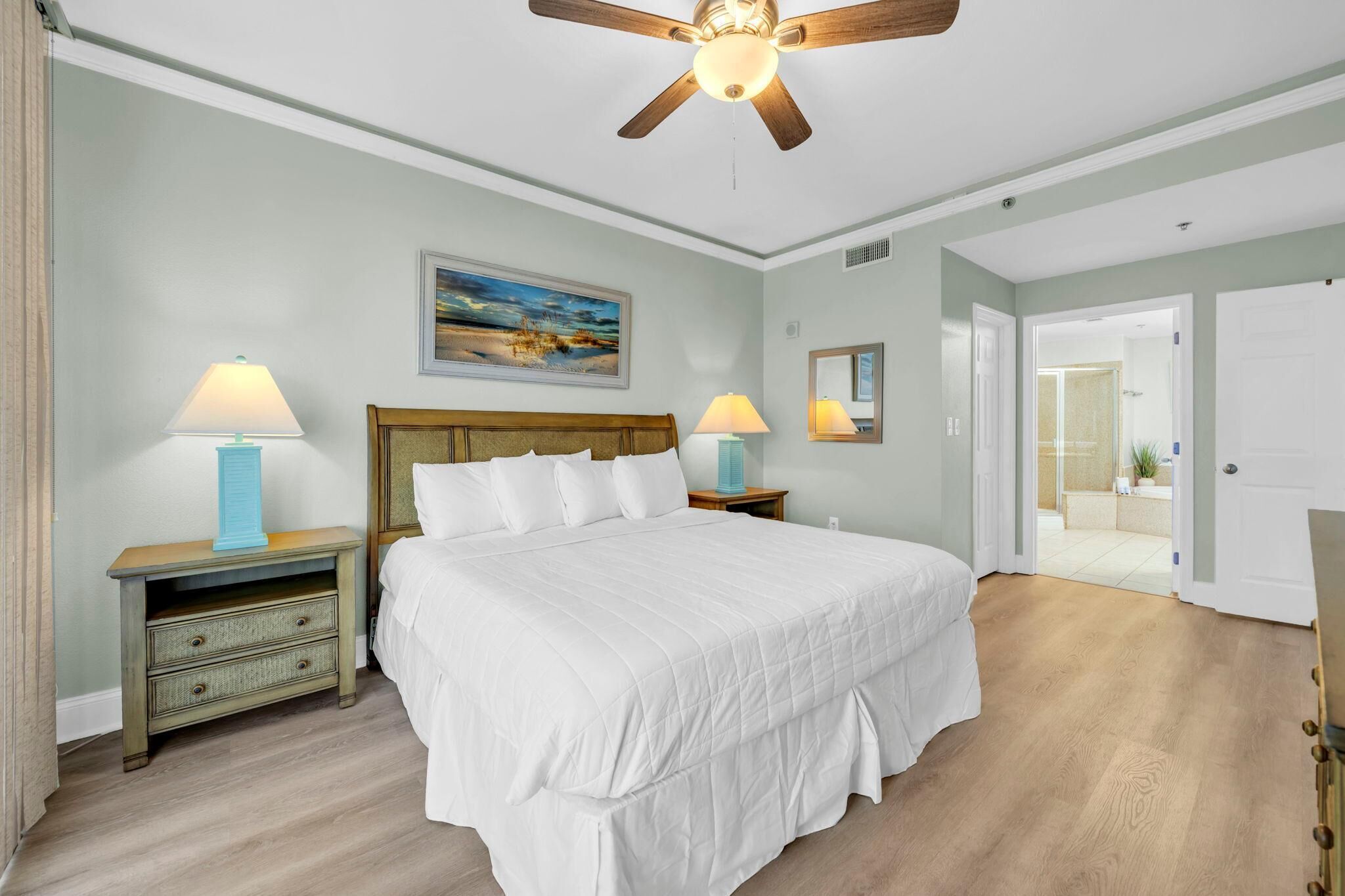 A bedroom with a bed, two bedside tables with lamps, a ceiling fan, a painting on the wall, and a door leading to another room.
