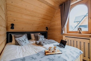 A cozy wooden cabin bedroom with a bed, two bedside lamps, a laptop, and a window with curtains.