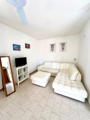 A white living room with a sectional sofa, a flat-screen TV on a white stand, and three framed artworks on the wall. - Living Terrace with Panoramic Views, 150 m from the Sea - Varco 2/3 Plemmirio (Punta Milocca)