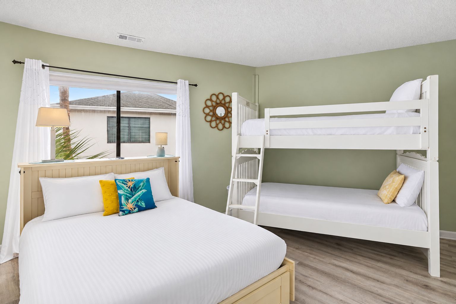 A room with a bunk bed, a single bed, a window with curtains, a lamp, and a wall decoration.