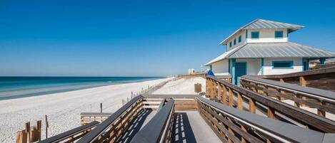 A beachside walkway leading to a building with a metal roof, wooden railings, and benches.