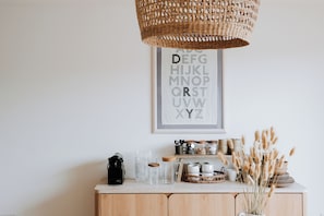 Un coin cuisine avec un meuble en bois, une machine à café, des bocaux en verre transparent et un luminaire suspendu en osier.