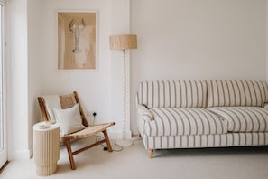 Un salon minimaliste avec un canapé à rayures, un fauteuil en osier, une lampe sur pied et une œuvre d’art encadrée accrochée au mur.