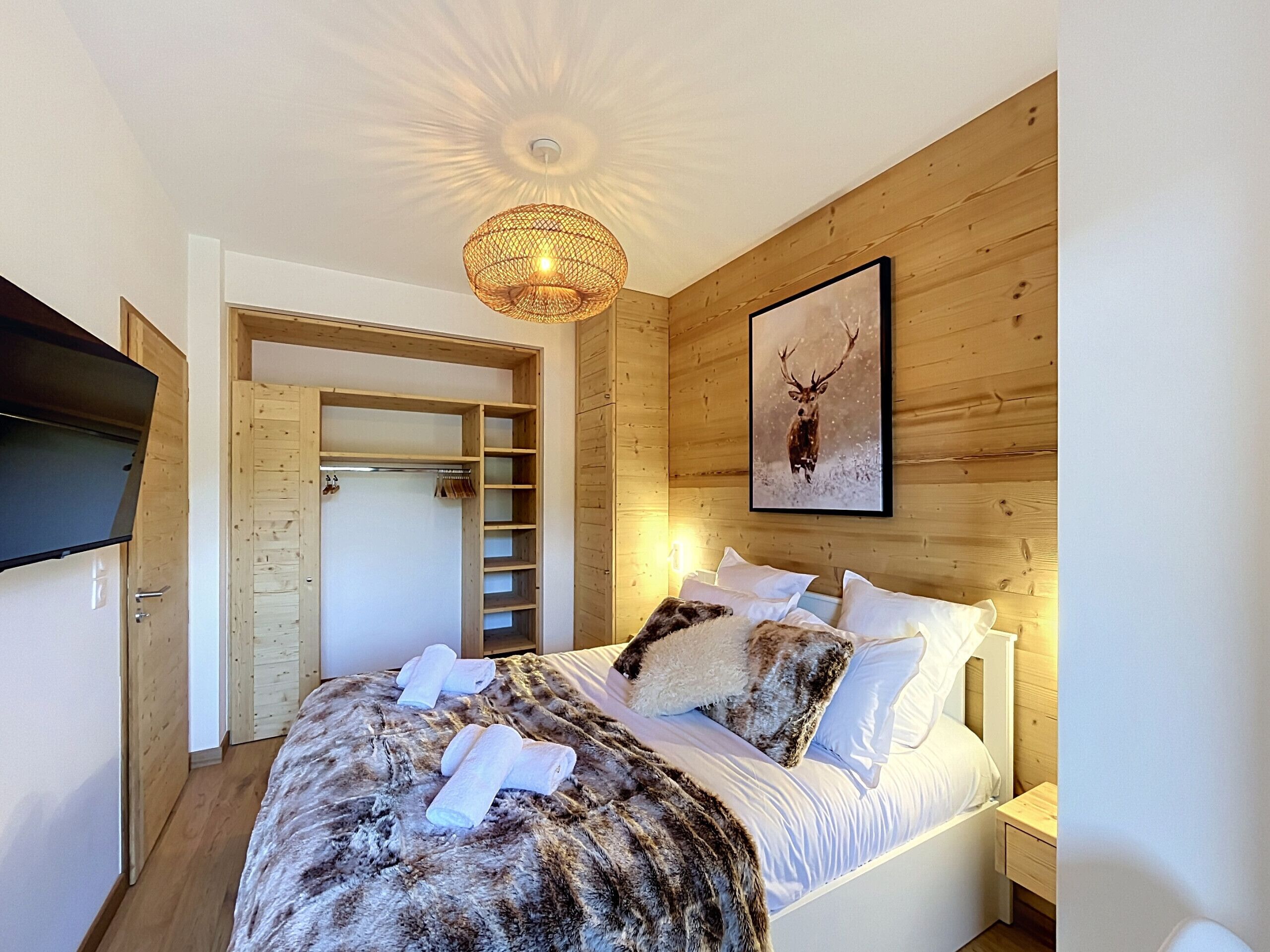 A bedroom with a bed, a wooden wardrobe, a television, a wall-mounted picture, and a ceiling light fixture.